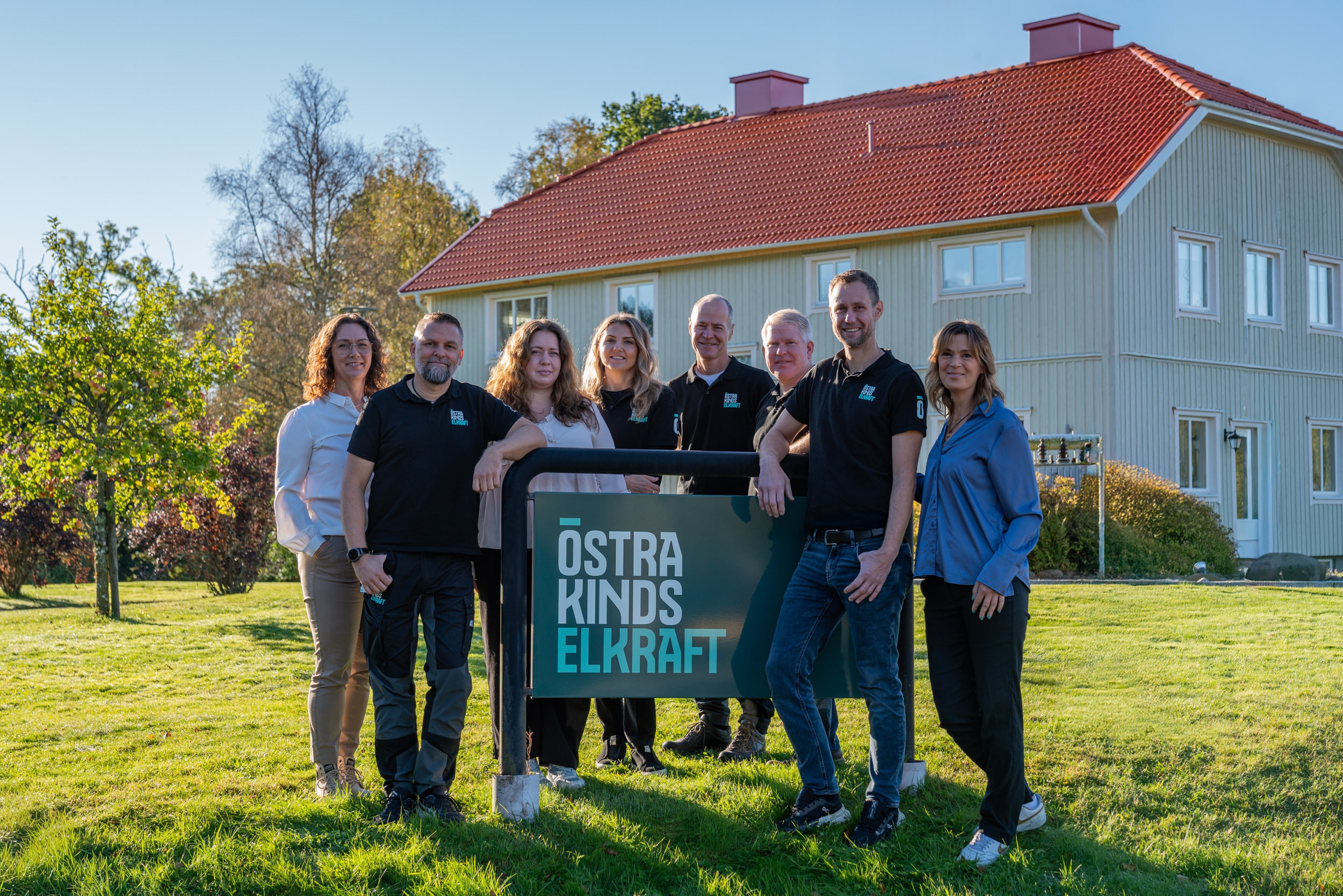 Gruppfoto av medarbetare som står utomhus vid en skylt med texten ”Östra Kinds Elkraft”, med en ljus träbyggnad och grön gräsmatta i bakgrunden i soligt väder.
